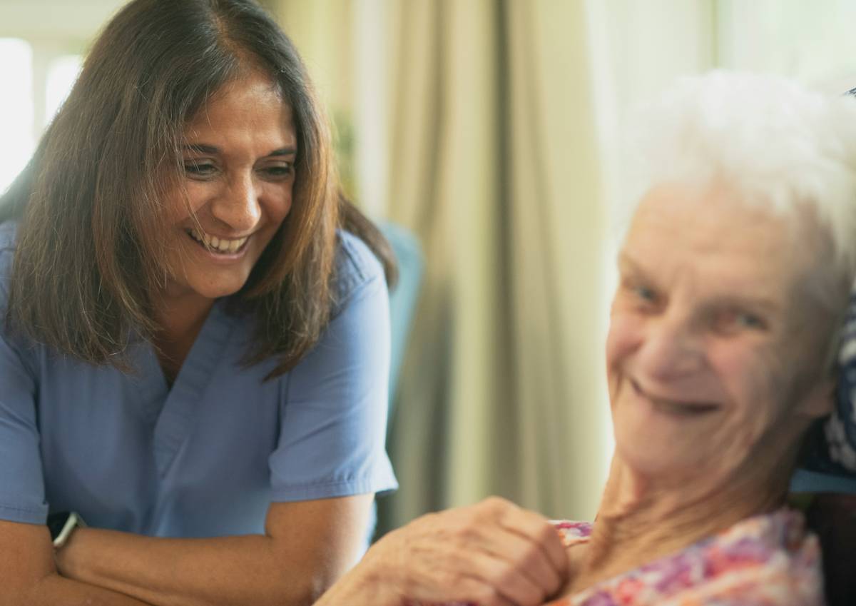 Senior woman smiling while seated in a sunlit common area with caregivers nearby — benefits of assisted living facilities